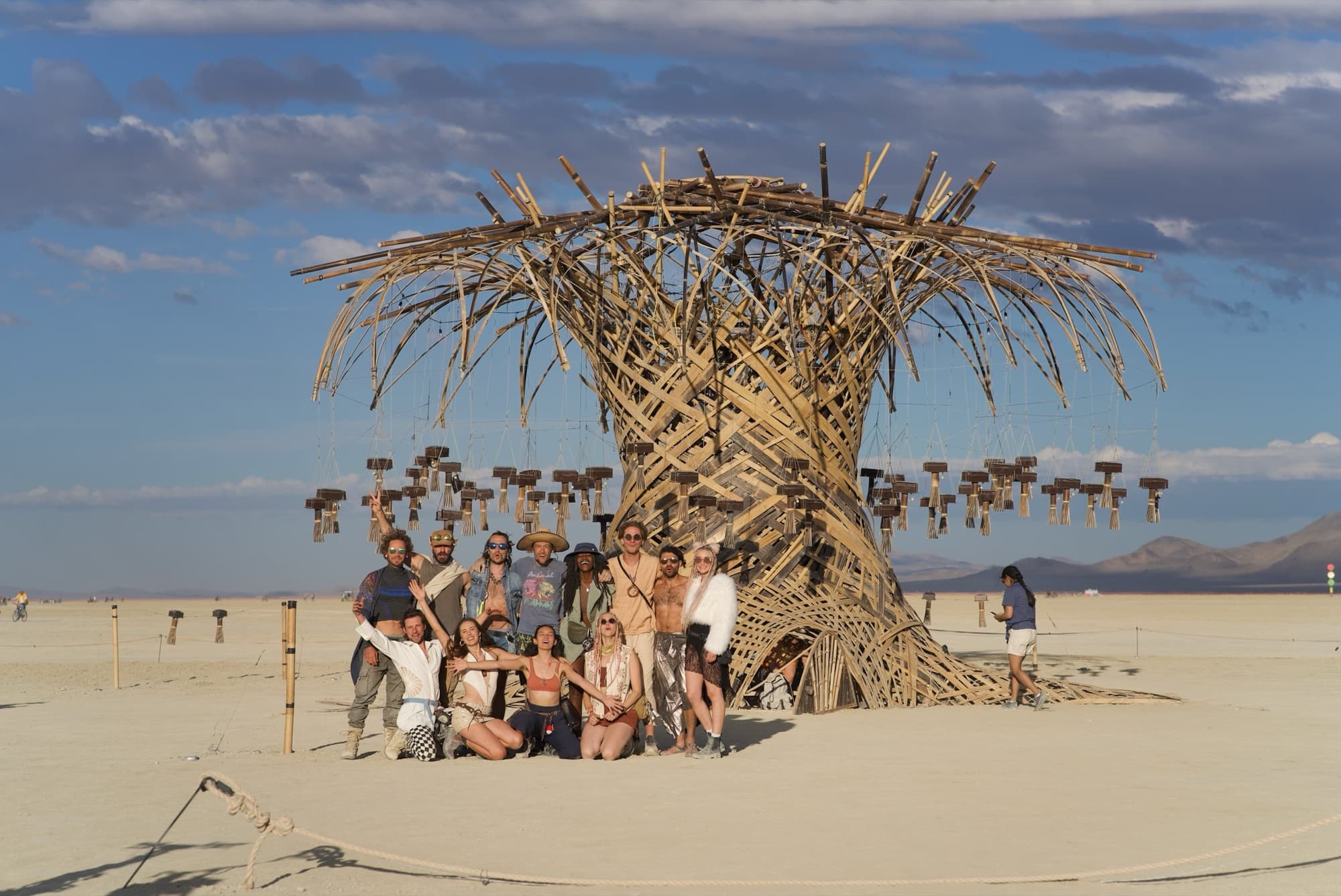 A group of about a dozen people gathered and posing at the foot of the finished Resonance Tree on the open playa under a blue sky