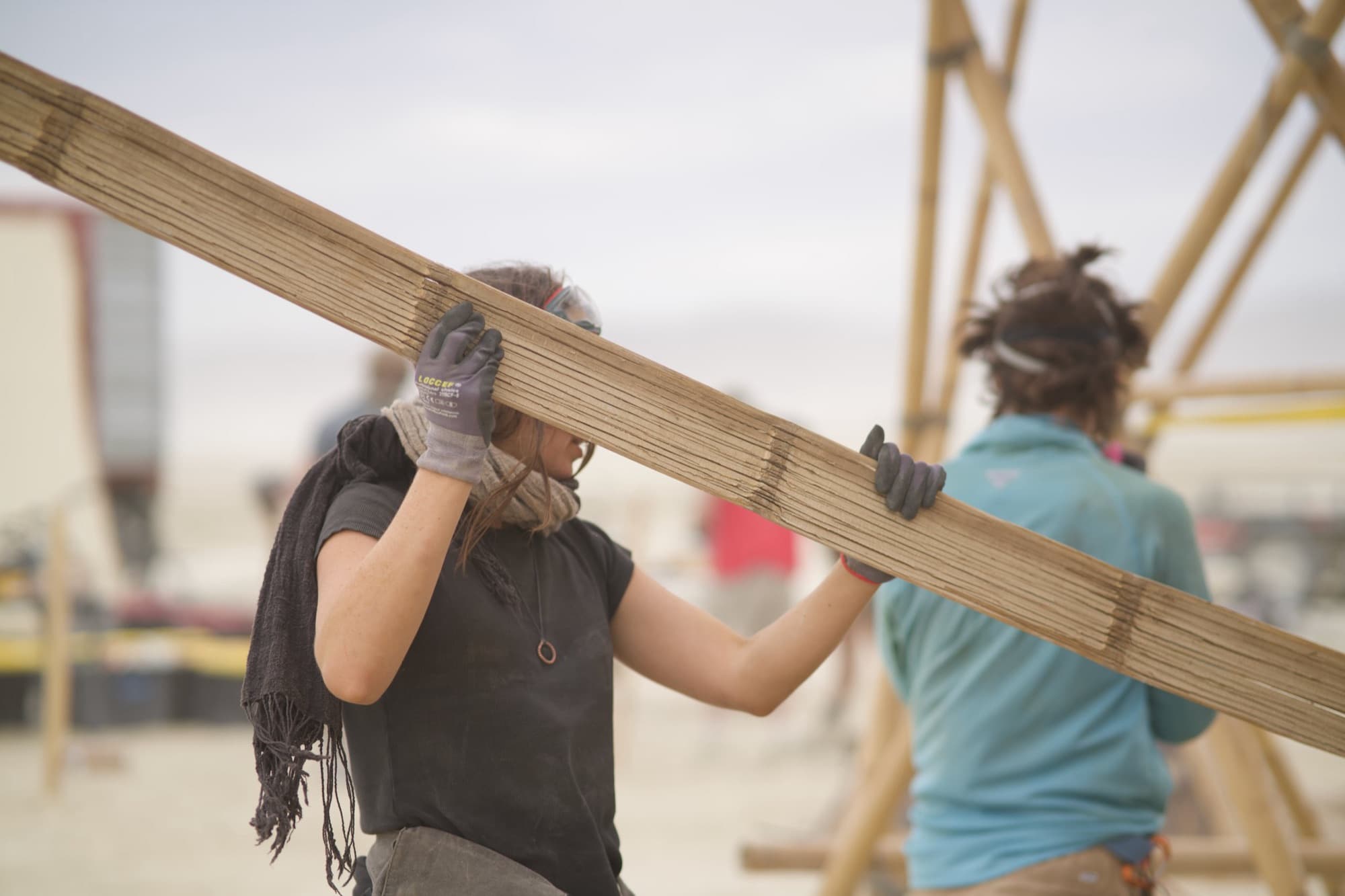 Two crew members carrying a long laminated bamboo member on their shoulders across the playa, the bamboo tower rising behind them