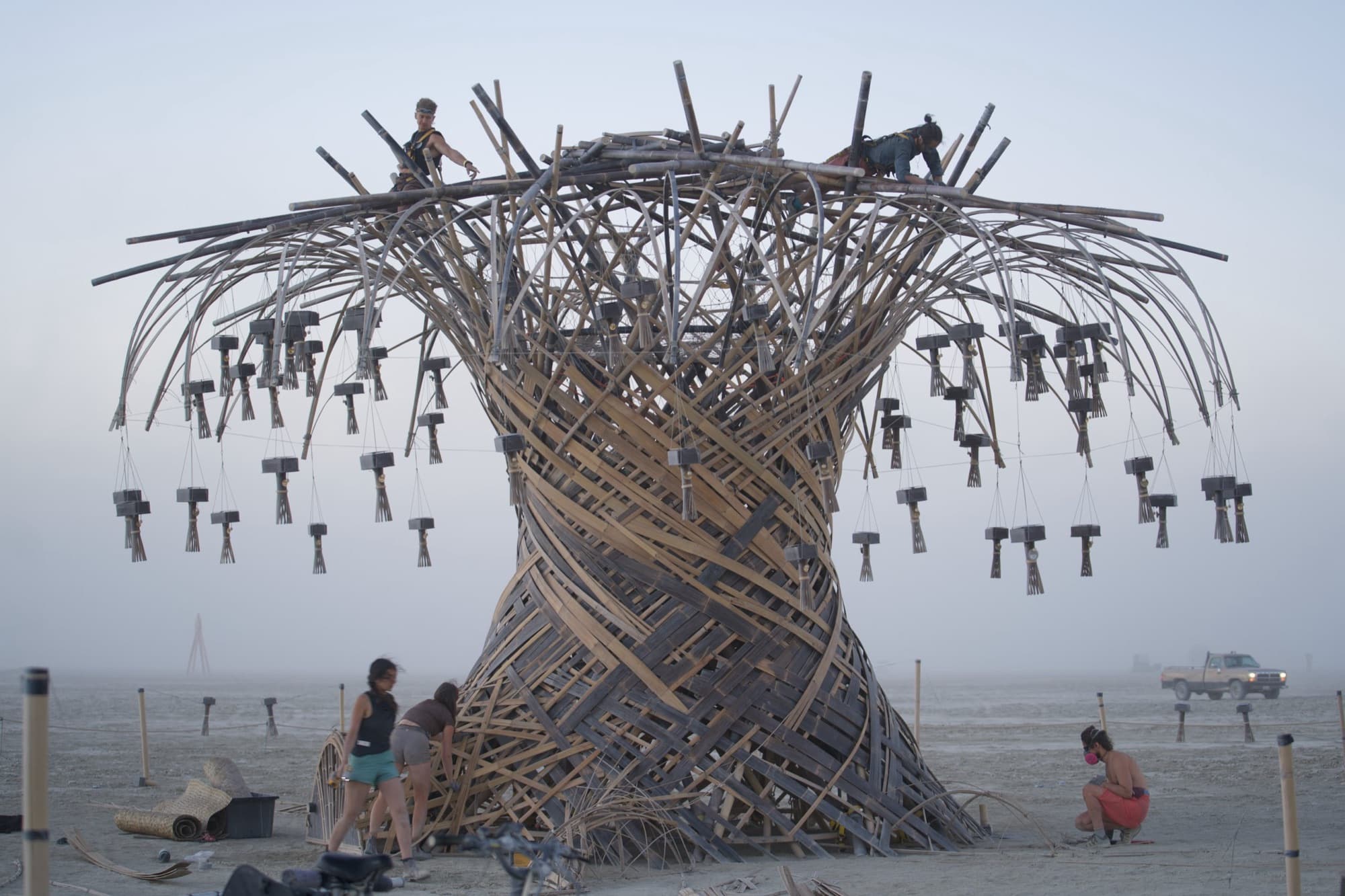 The finished Resonance Tree head-on at dusk with its chimes hung, two crew on the crown and visitors at the base
