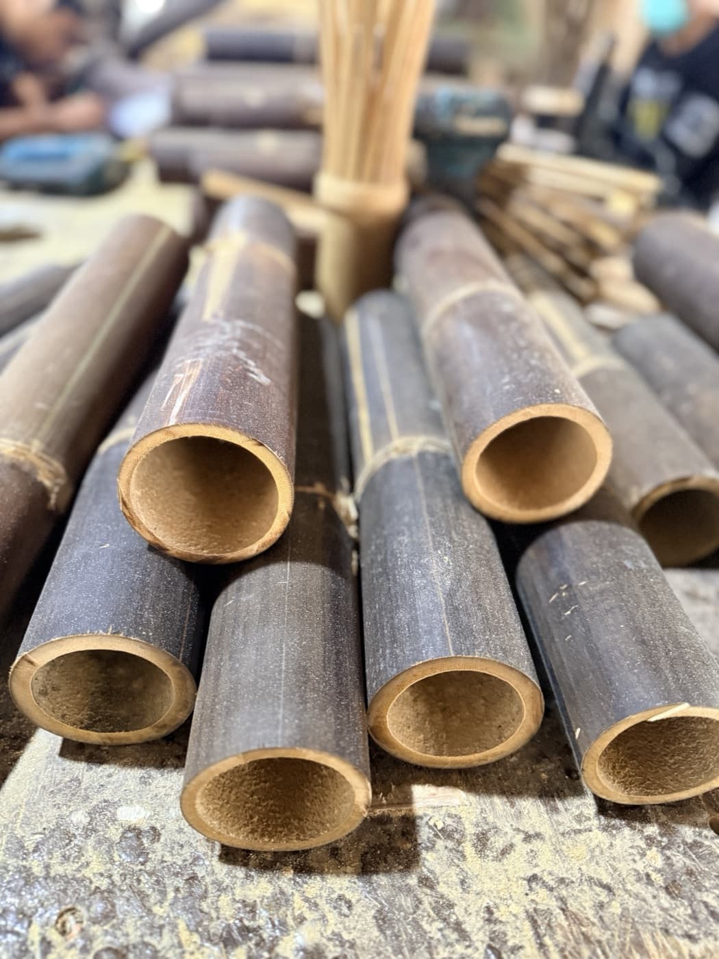 Cross-cut ends of dark treated Petung bamboo poles stacked on a workshop bench, their hollow interiors and wall thickness facing the camera