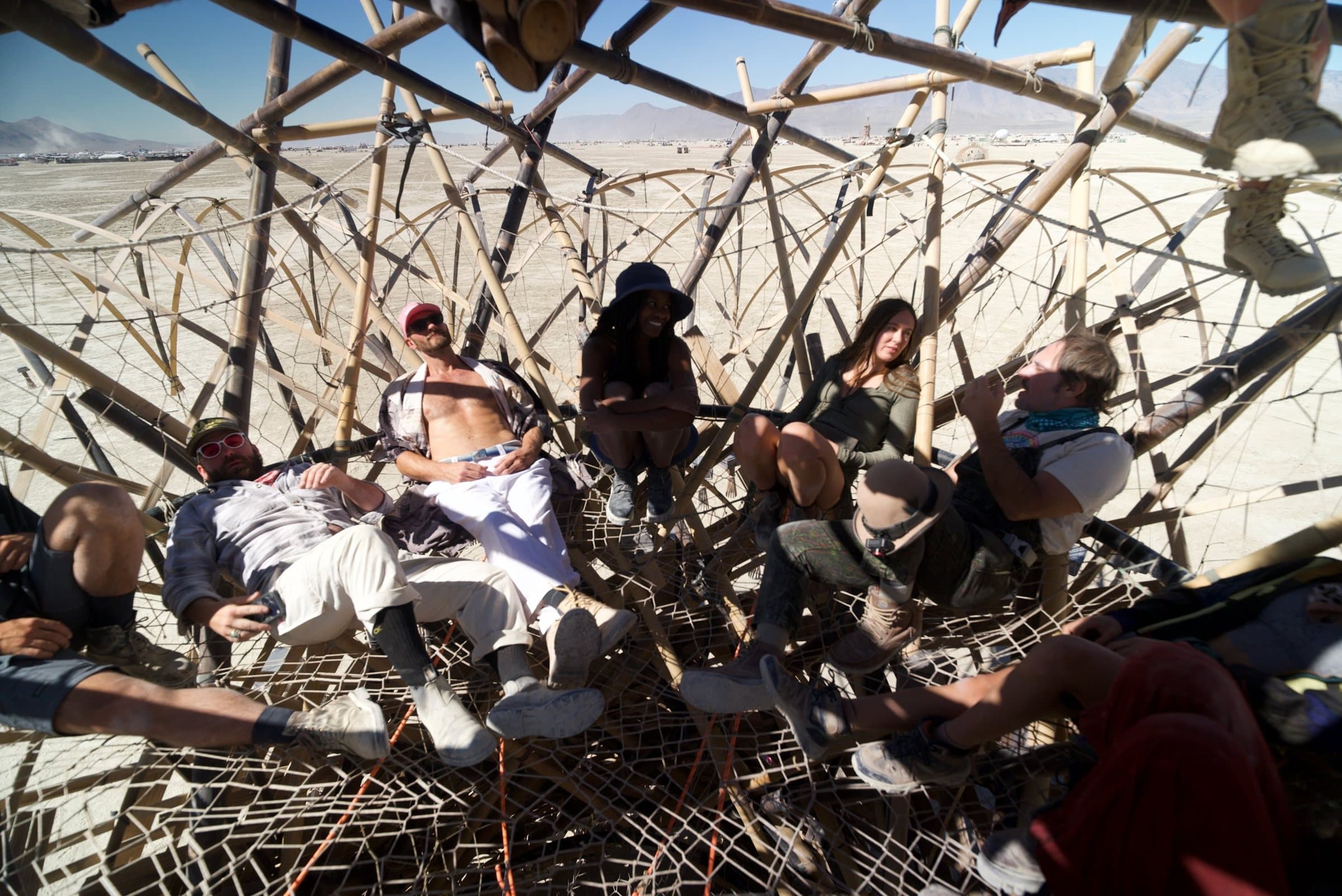 The crew lounging in the rope nets inside the canopy at midday, the bamboo web crossing overhead and the playa beyond