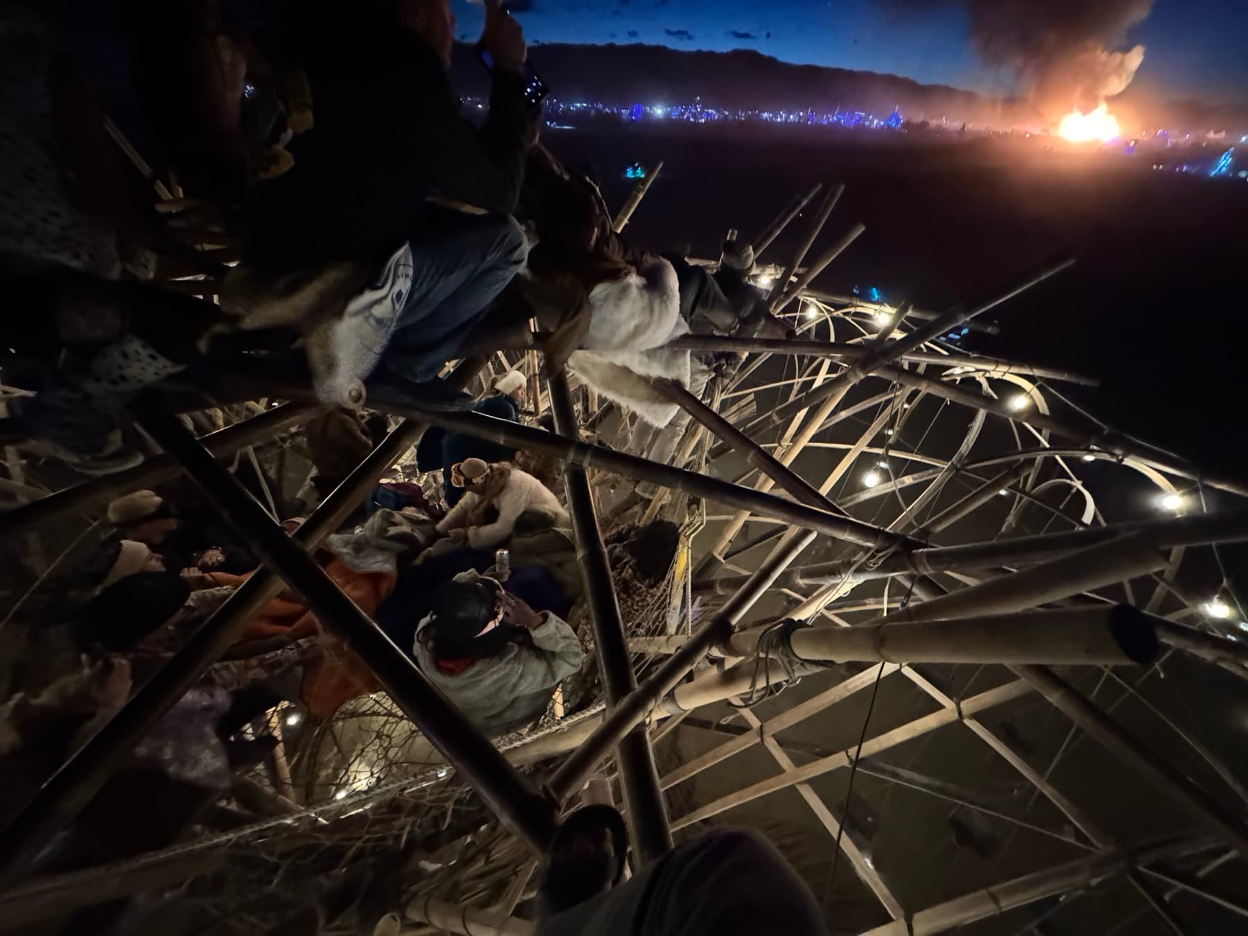 Looking down into the Resonance Tree's crown at night: people bundled in blankets and furs across the woven nets, with the lit city and a distant fire on the horizon