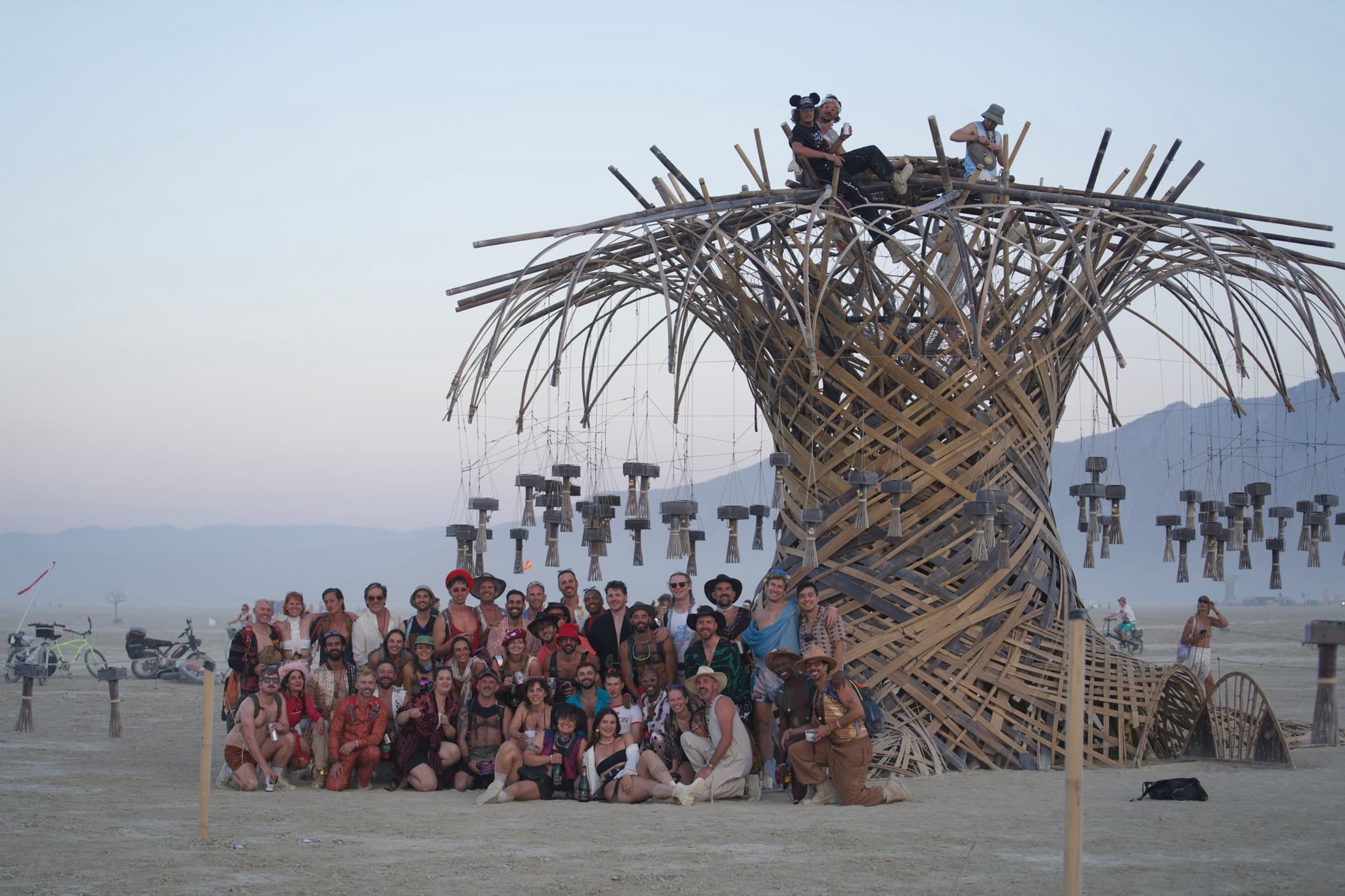 Roughly four dozen crew members gathered and seated at the foot of the finished Resonance Tree at dusk, two of them up on the crown, the desert and mountains beyond