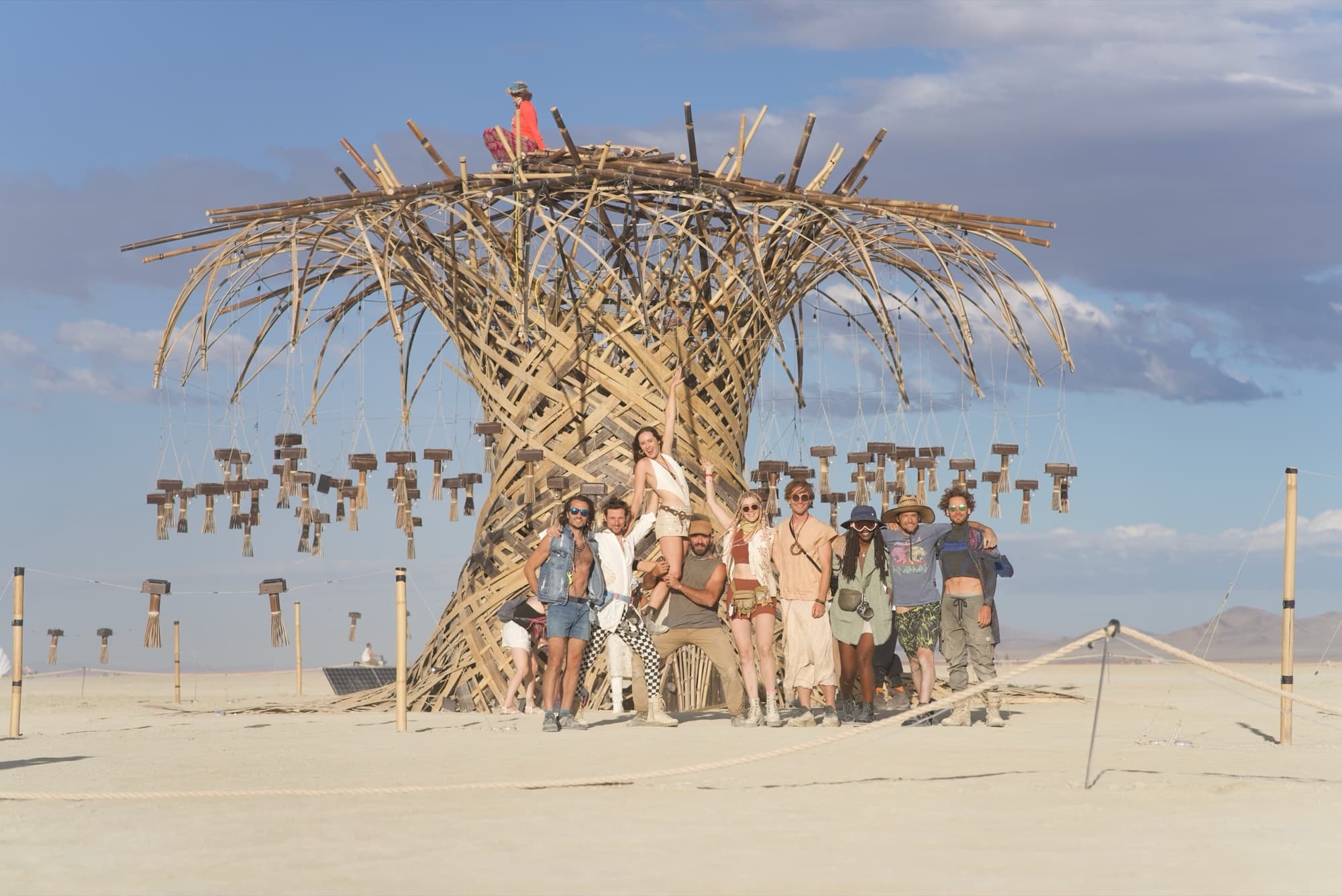 A crowd of visitors gathered under the Resonance Tree on the playa, one person up on the crown