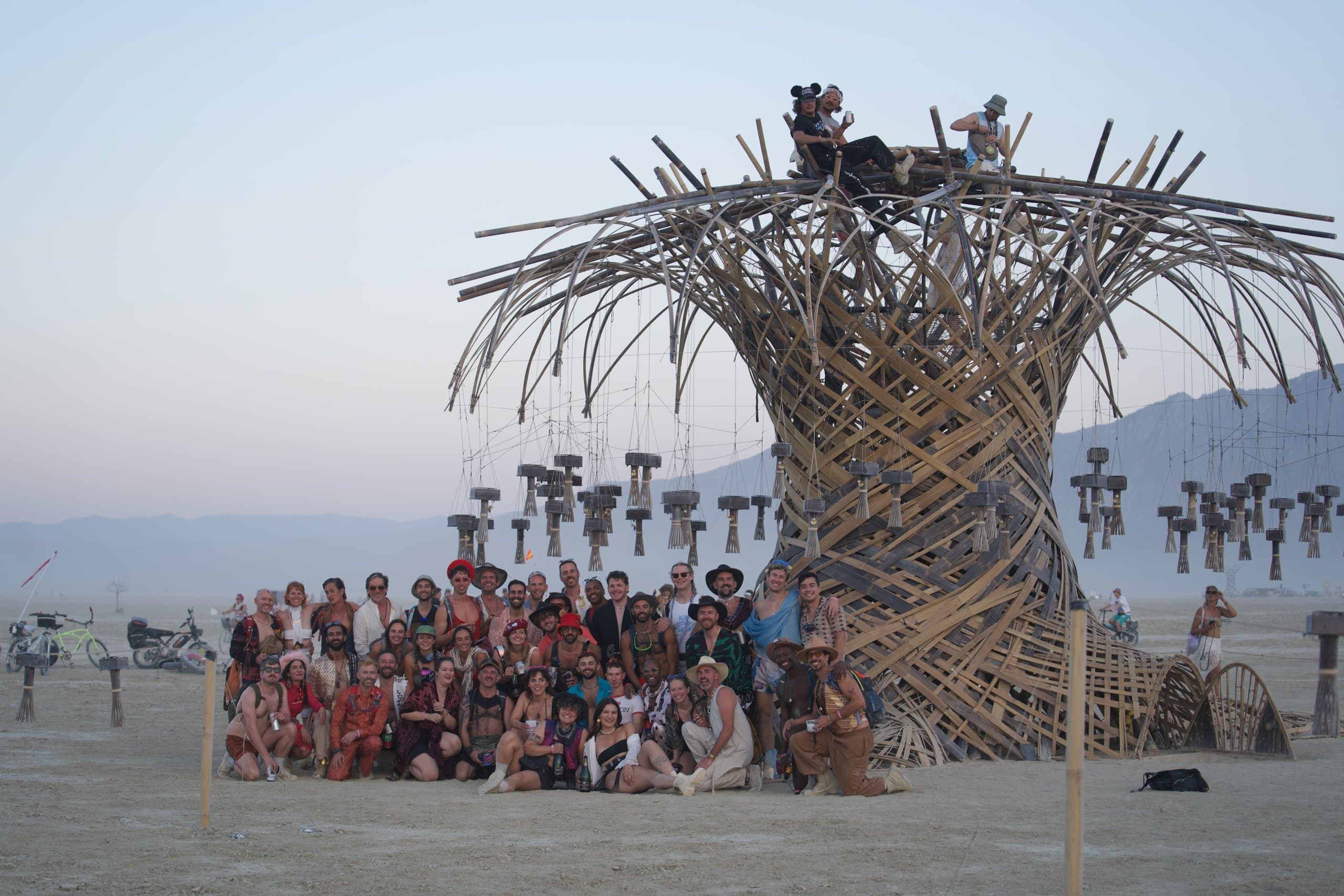 The whole build crew gathered in front of the finished Resonance Tree at golden hour, chimes hanging above them