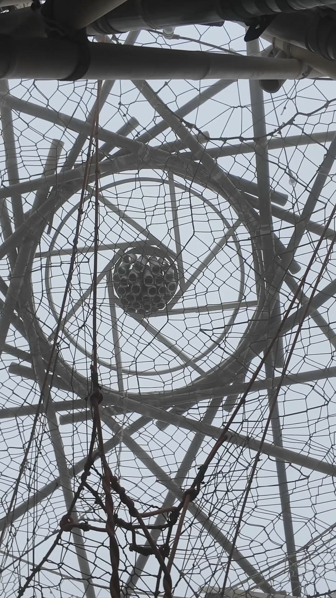 Looking straight up into the woven bamboo crown of the Resonance Tree: rings, nets and the pole hub against a pale sky