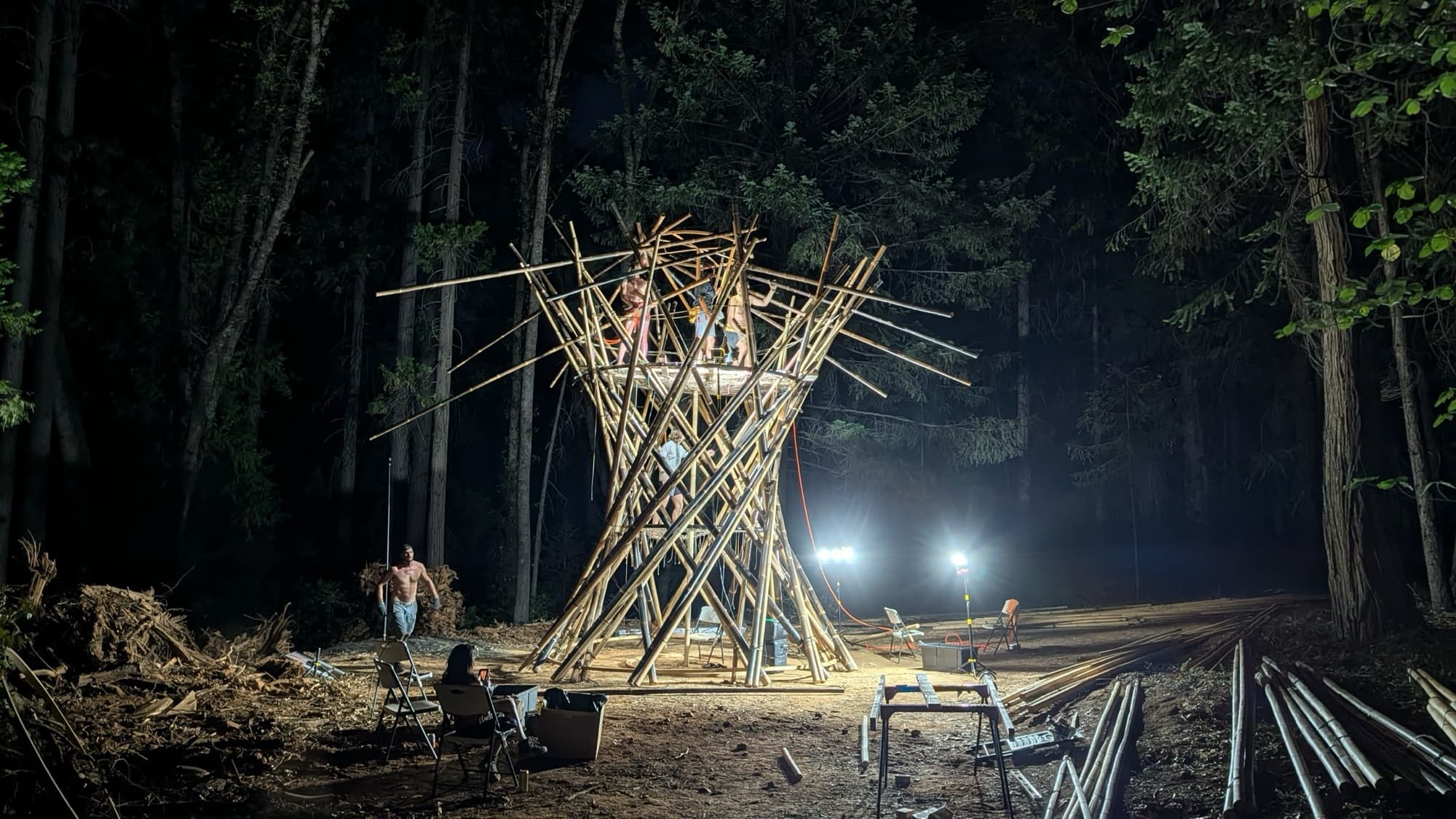 The bamboo trunk lattice lit from below at night during its Nevada City test raise, work lights glowing between the pines and poles stacked beside it