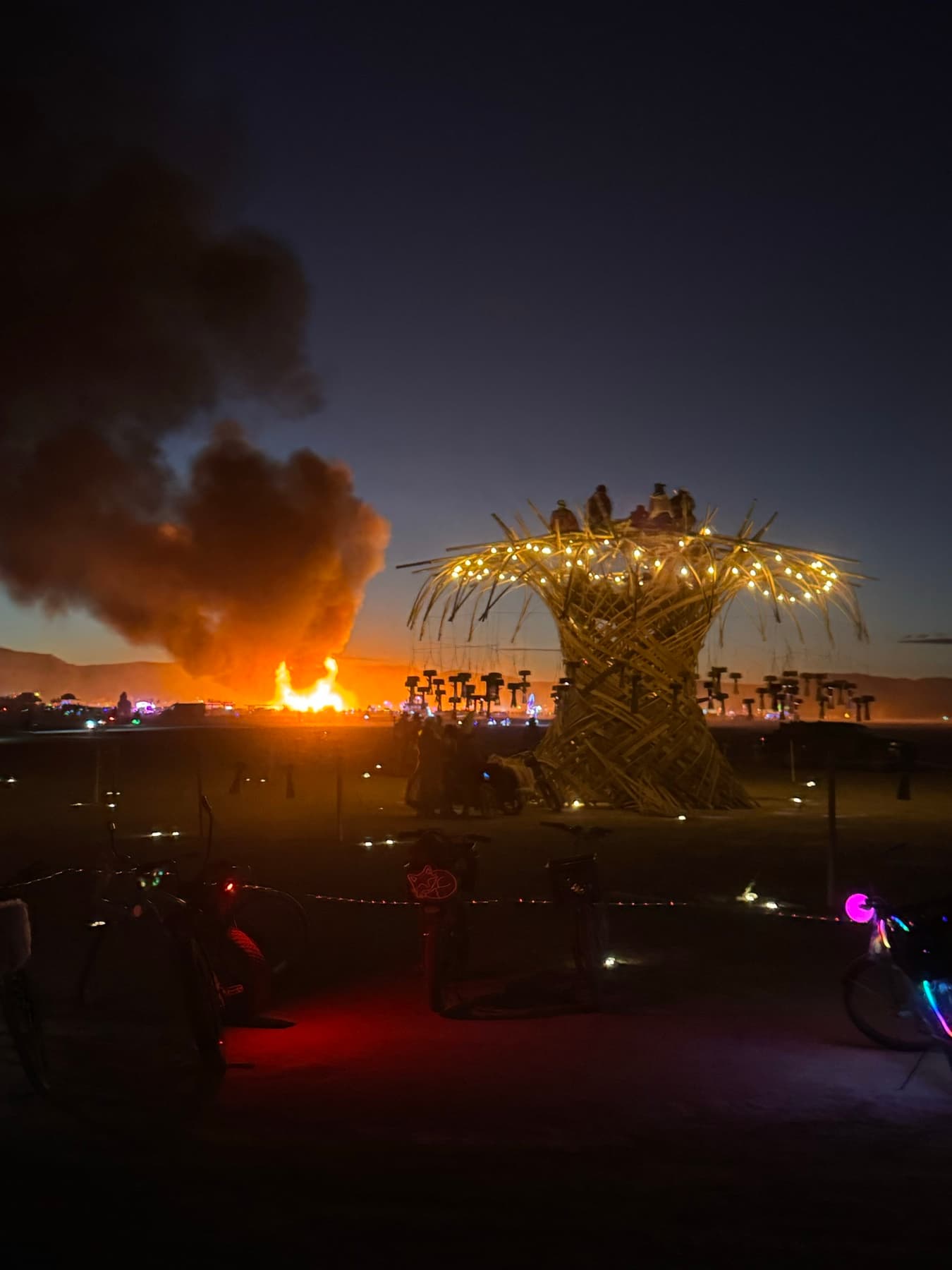 The lit Resonance Tree at night with visitors on its crown, bikes parked at its roots, and the burn blazing on the horizon behind it