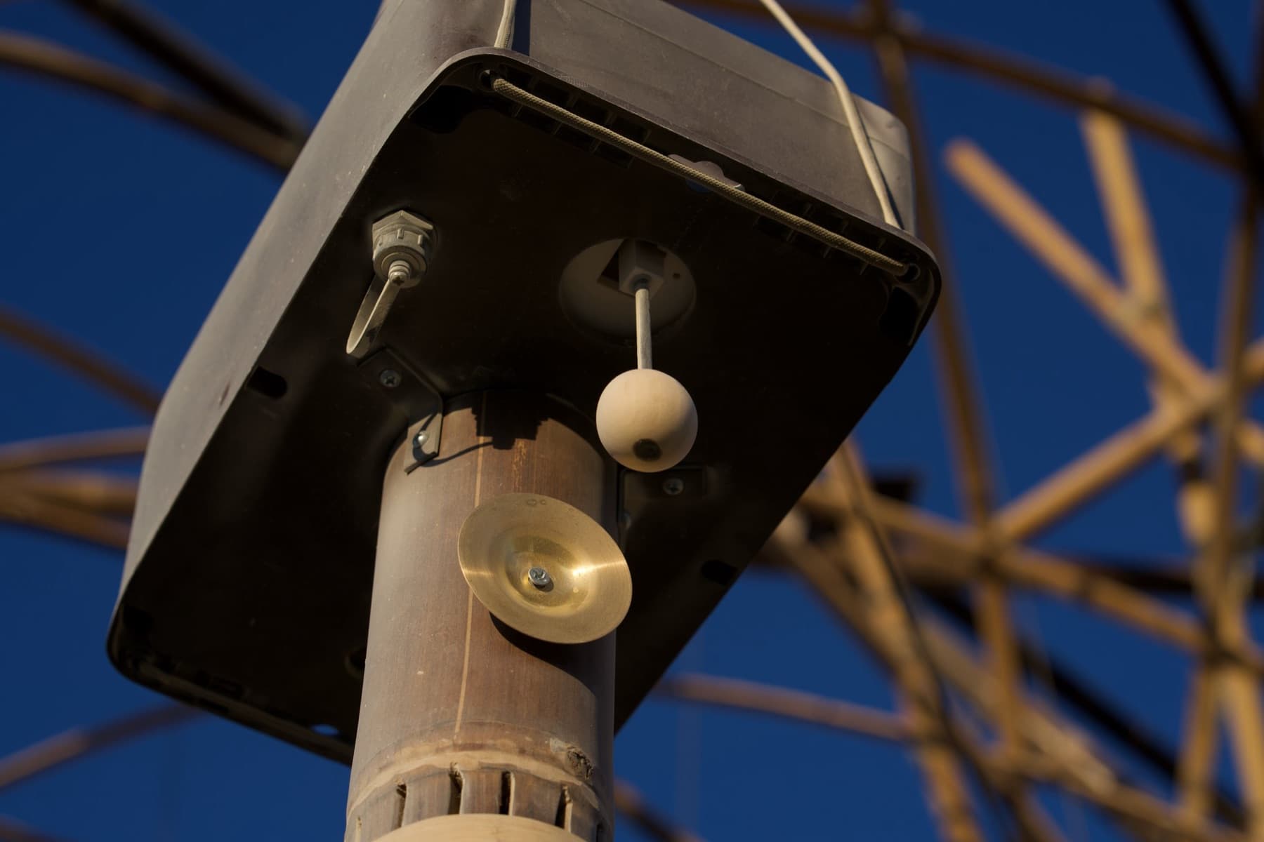 Close-up of a single bamboo chime showing the solar head, the striker ball on its arm, and the brass disc against a blue sky