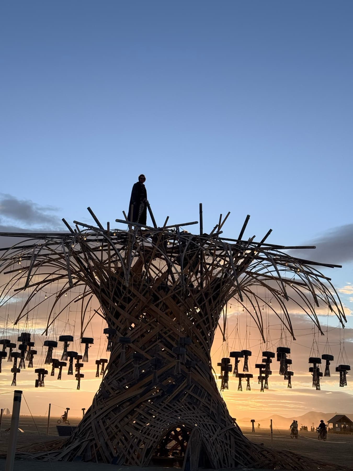 A lone figure standing on the crown of the Resonance Tree at sunrise, hanging chimes silhouetted against the glowing horizon