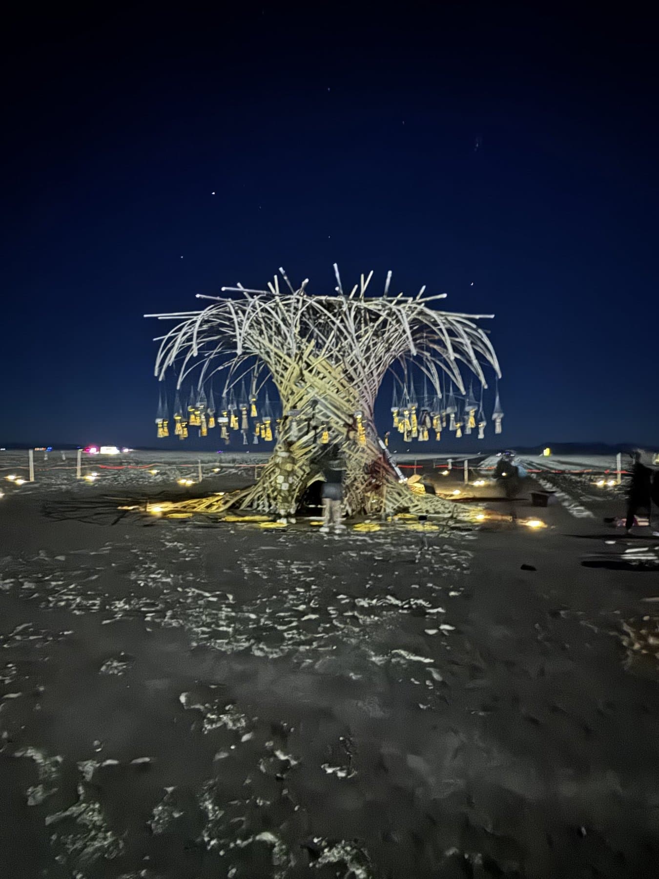 The Resonance Tree lit from within at night, lanterns glowing and blue light pooled across the playa around its base