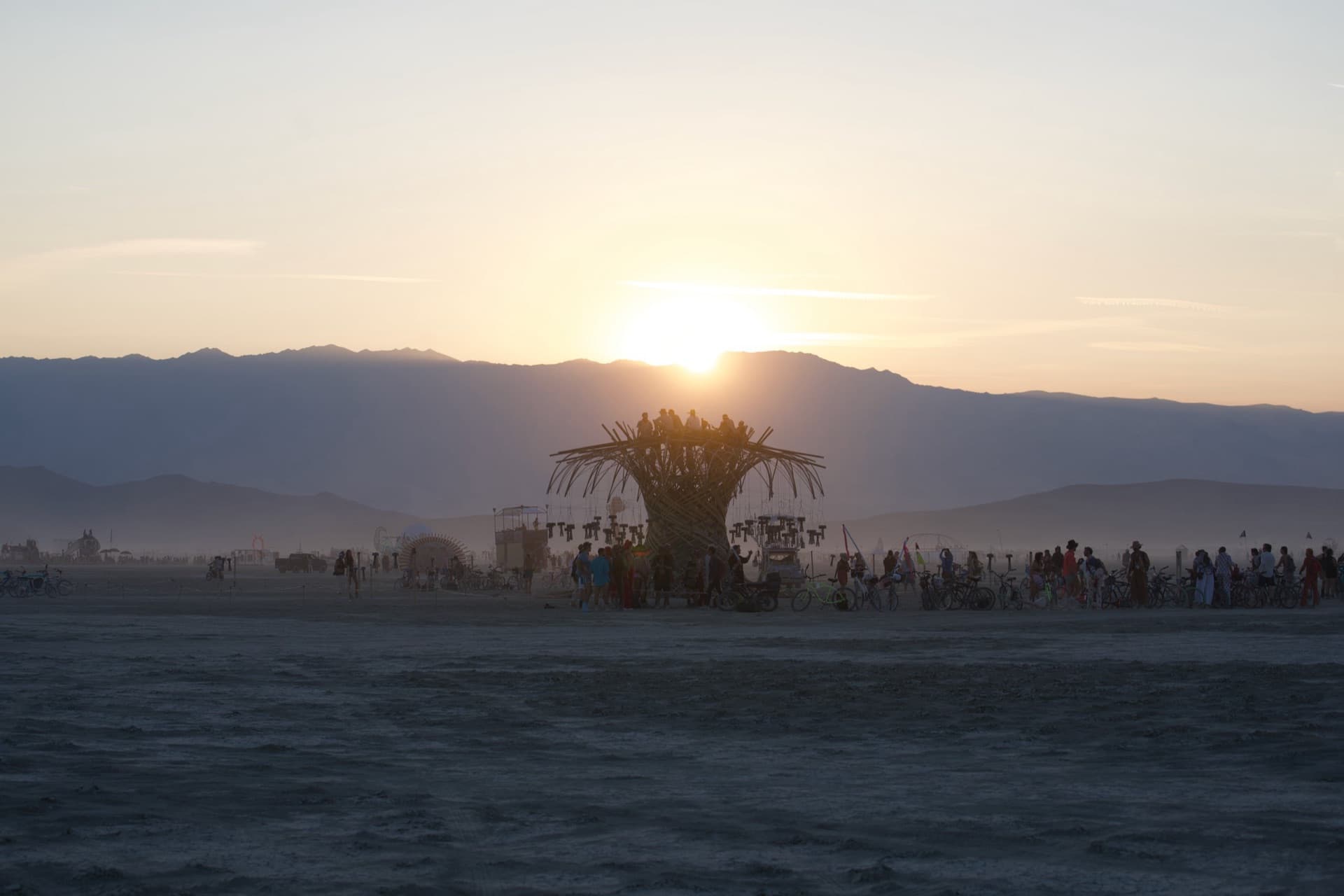 Sunset on the horizon behind the Resonance Tree, the playa crowd drifting toward it