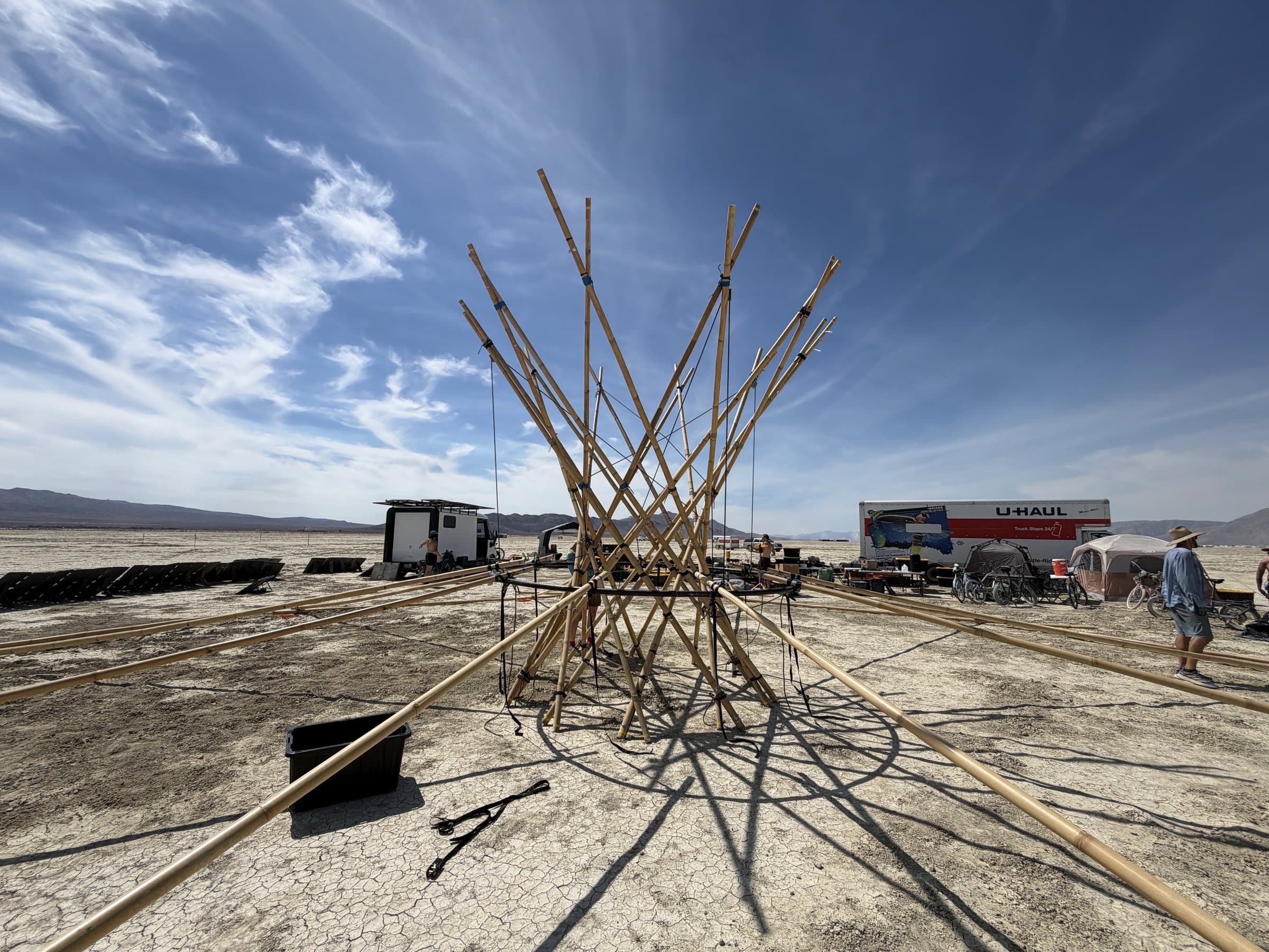 The first bamboo of the Resonance Tree raised on the playa: the lashed cone from a low angle under a wide sky, the crew's camp behind