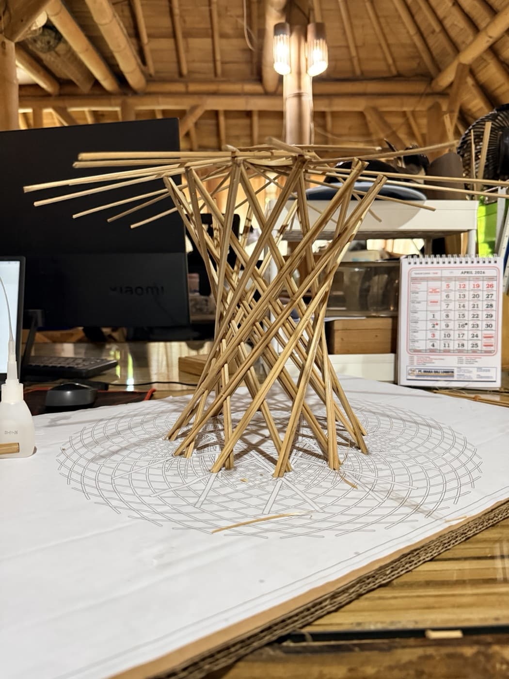 A bamboo-skewer scale model of the Resonance Tree standing on its printed plan drawing in the workshop, the X-frame trunk flaring into a splayed canopy