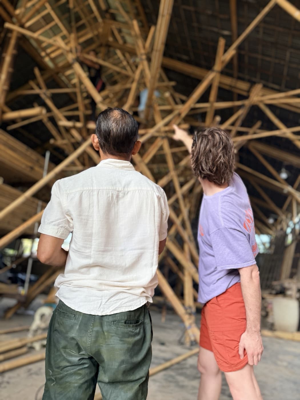 Two people seen from behind looking up into the dense bamboo gridshell of the trunk in the workshop, one pointing up into the weave